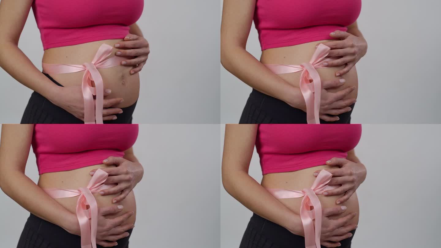 特写镜头。一个肚子上系着粉色丝带的孕妇站在白色背景上。一个女人温柔地用双手抱住自己的肚子。