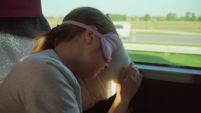 戴着睡眠面罩的女孩在高速公路上的长途旅行中倚靠在车窗上休息。关闭了。疲惫的孩子在巴士上戴着眼罩遮挡阳