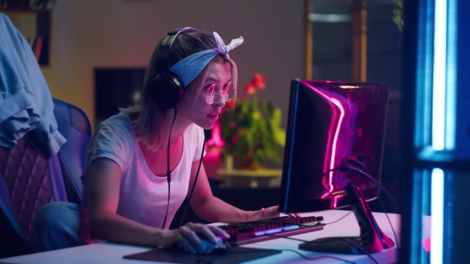戴着耳机玩电脑游戏的年轻女子，赢家。女玩家戴着眼镜，看着电脑显示器，使用电脑鼠标和键盘，赢得了艰苦的