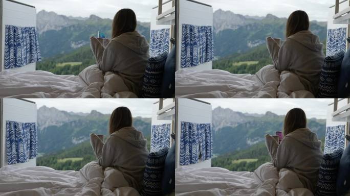 年轻舒适的女性在露营车的床上浏览电话，早上喝咖啡，欣赏风景秀丽的山景