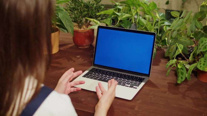 花店女用笔记本电脑视频通话，蓝色度键屏。有虚拟会议的温室工人，女植物学家，小企业主。园丁在花店，植物