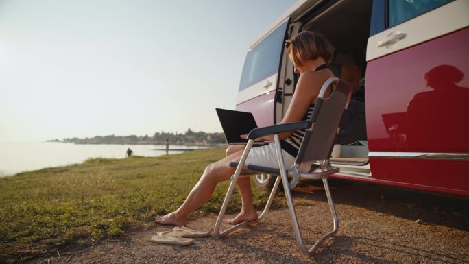 微笑的女性自由职业者在露营车外的椅子上用笔记本电脑工作的SLO镜头