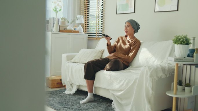 放松的亚洲癌症患者戴着头巾享受她的闲暇时间看电视，坐在舒适的沙发在明亮的生活空间。与癌症共存。