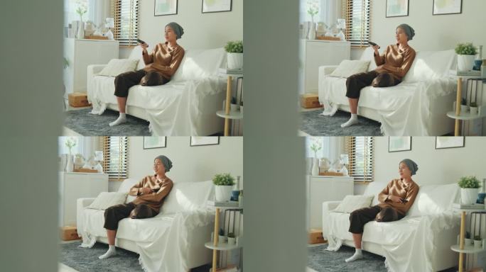 放松的亚洲癌症患者戴着头巾享受她的闲暇时间看电视，坐在舒适的沙发在明亮的生活空间。与癌症共存。