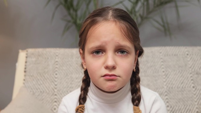 小女孩不开心穿着白色高领毛衣扎着小辫儿坐在沙发上对着镜头生气准备哭被冒犯了