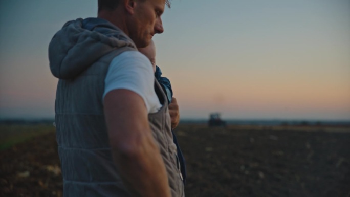 日落时分，男子抱着男婴站在农场边玩手机