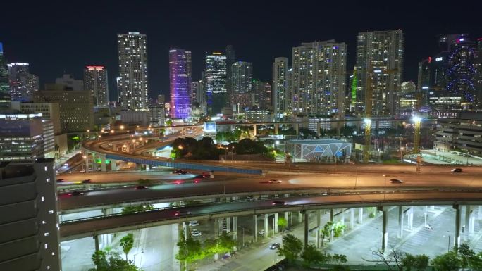 位于佛罗里达州迈阿密的一座现代美国城市，夜间高速公路交叉口上方有高层办公楼，车辆疾驶。美国城市基础设