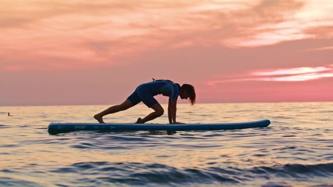 在日落的海洋中，健身女性在冲浪板上练习瑜伽时伸展的慢镜头