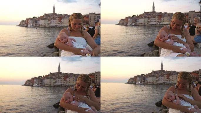 夕阳下，快乐的母亲抱着蹒跚学步的孩子坐在海边的岩石上。罗维尼，伊斯特拉，克罗地亚。