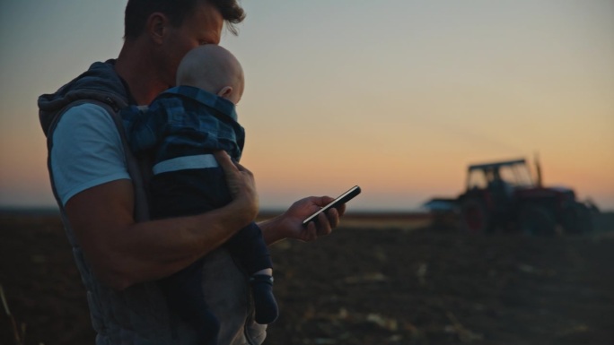 SLO MO男农民带着儿子在日落时分用手机站在农场