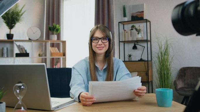 戴着耳机微笑的年轻女子坐在摄像机前，准备好了有关她工作的文件