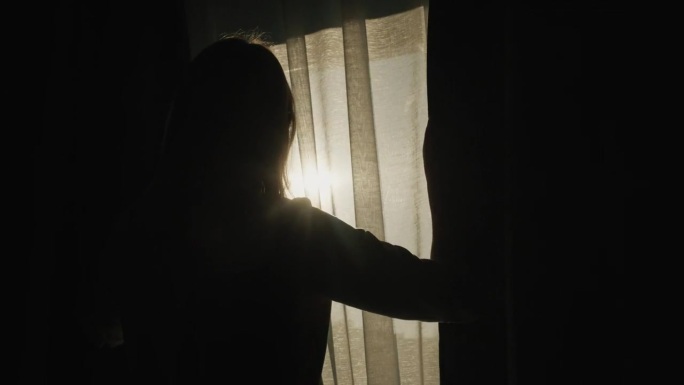 一个拉开遮光窗帘的女人的剪影。它被从窗户照进来的明亮的阳光照亮了