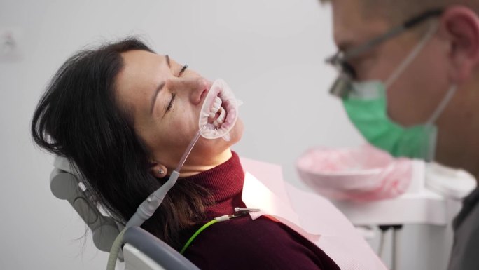 现代牙科诊所中，男牙医用双筒望远镜治疗女病人的牙齿。在安装贴面之前，医生用注射器将蓝色蚀刻凝胶涂在牙