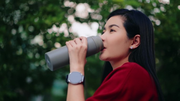 在以树木为背景的公园里，一名身穿红色休闲服、戴着智能手表的亚洲女性正在用灰色的保温瓶喝水。生活平衡和