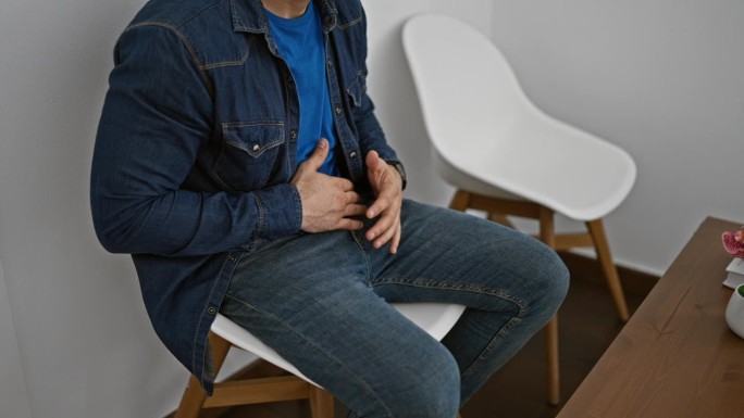 一名忧心忡忡的西班牙裔年轻男子在阳光明媚的候诊室里休息，坐在舒适的椅子上抚摸着疼痛的肚子，看上去严肃