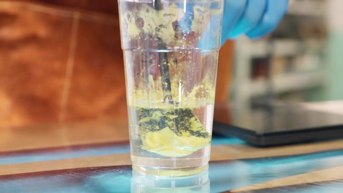 时尚的透明环氧树脂在塑料杯中与黄色颜料混合，形成多组分溶液，用于涂覆单个木制桌面。工业用混合器搅拌两