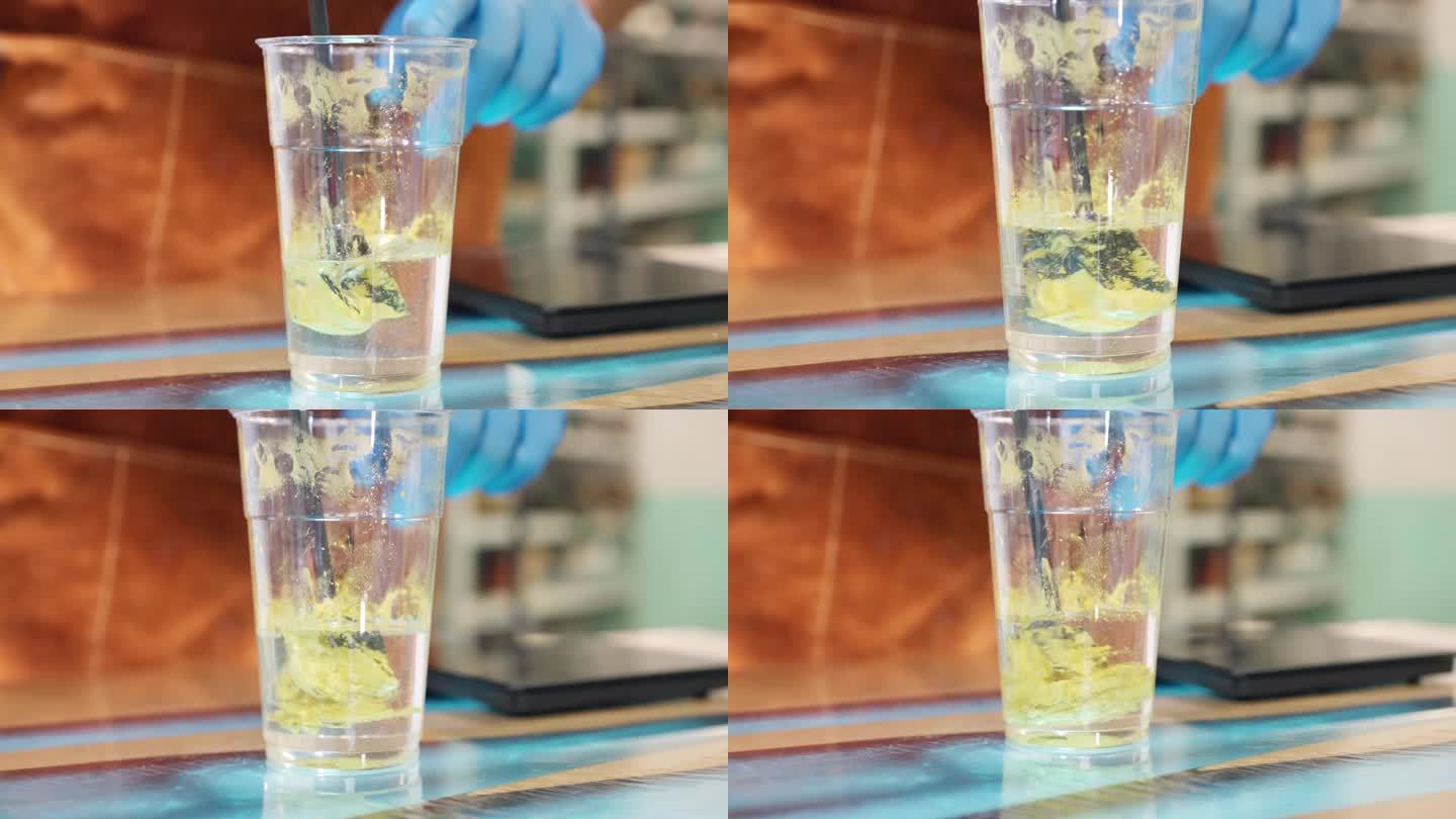 时尚的透明环氧树脂在塑料杯中与黄色颜料混合，形成多组分溶液，用于涂覆单个木制桌面。工业用混合器搅拌两
