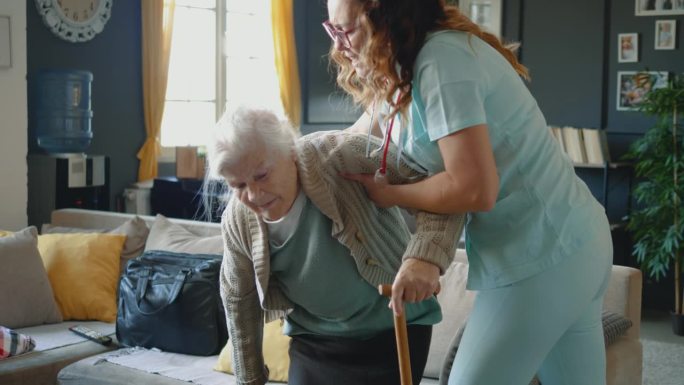 协助高龄祖母在家行走的护士。一位老妇人的腿有问题，走路困难，在看护人的帮助下站起来时感到头晕