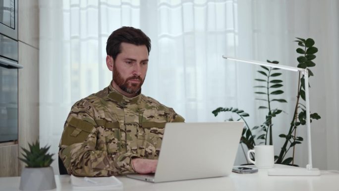 自信的美军士兵坐在家里的工作场所，用无线笔记本电脑打字。身穿军装的大胡子男子表情严肃地看着镜头。