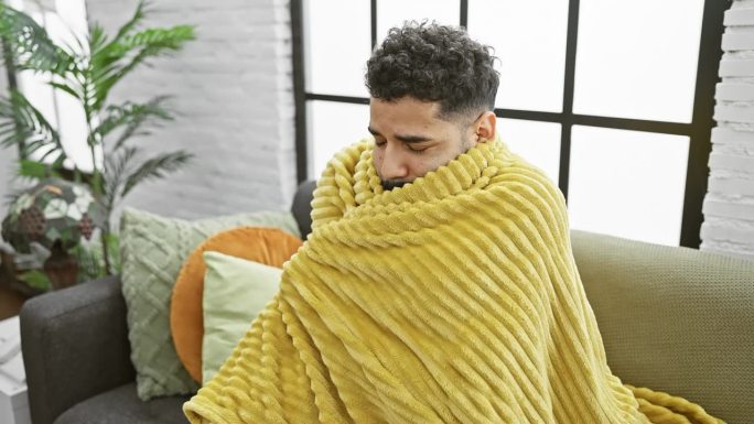 一个裹着黄毯子的年轻人若有所思地坐在摆满植物和靠垫的舒适室内。