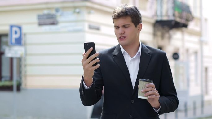 一个年轻的商人走在街上，正在进行视频通话。穿着经典西装的人，一杯咖啡