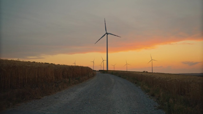 通往明天的道路:黄昏时分，麦田里的土路通往旋转的风力涡轮机