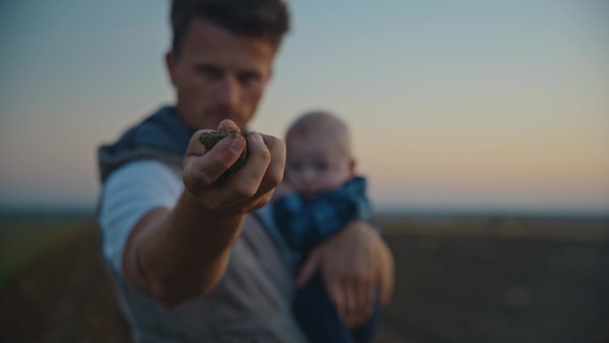 日落时分，男农艺师抱着儿子在田间播种前检查土壤质量的锁定镜头