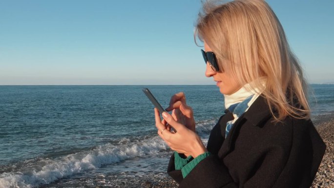 年轻女子在大海或海洋背景下使用智能手机。穿大衣走路。