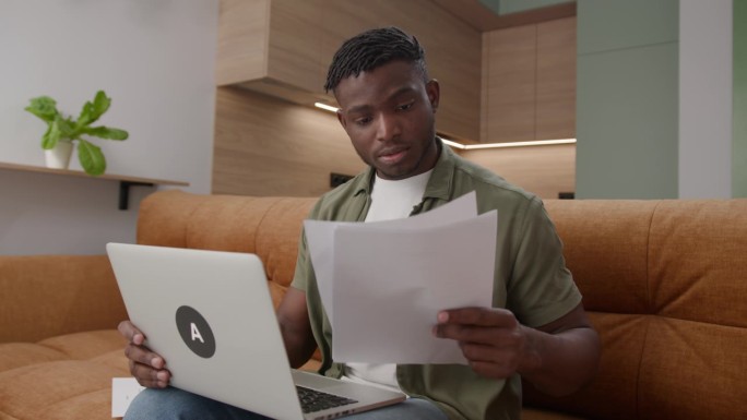 用笔记本电脑处理文件的黑人。30多岁的企业家坐在沙发上管理文件、发票、税单。