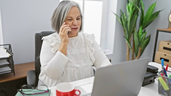Senior woman conversing on phone at desk with lapt