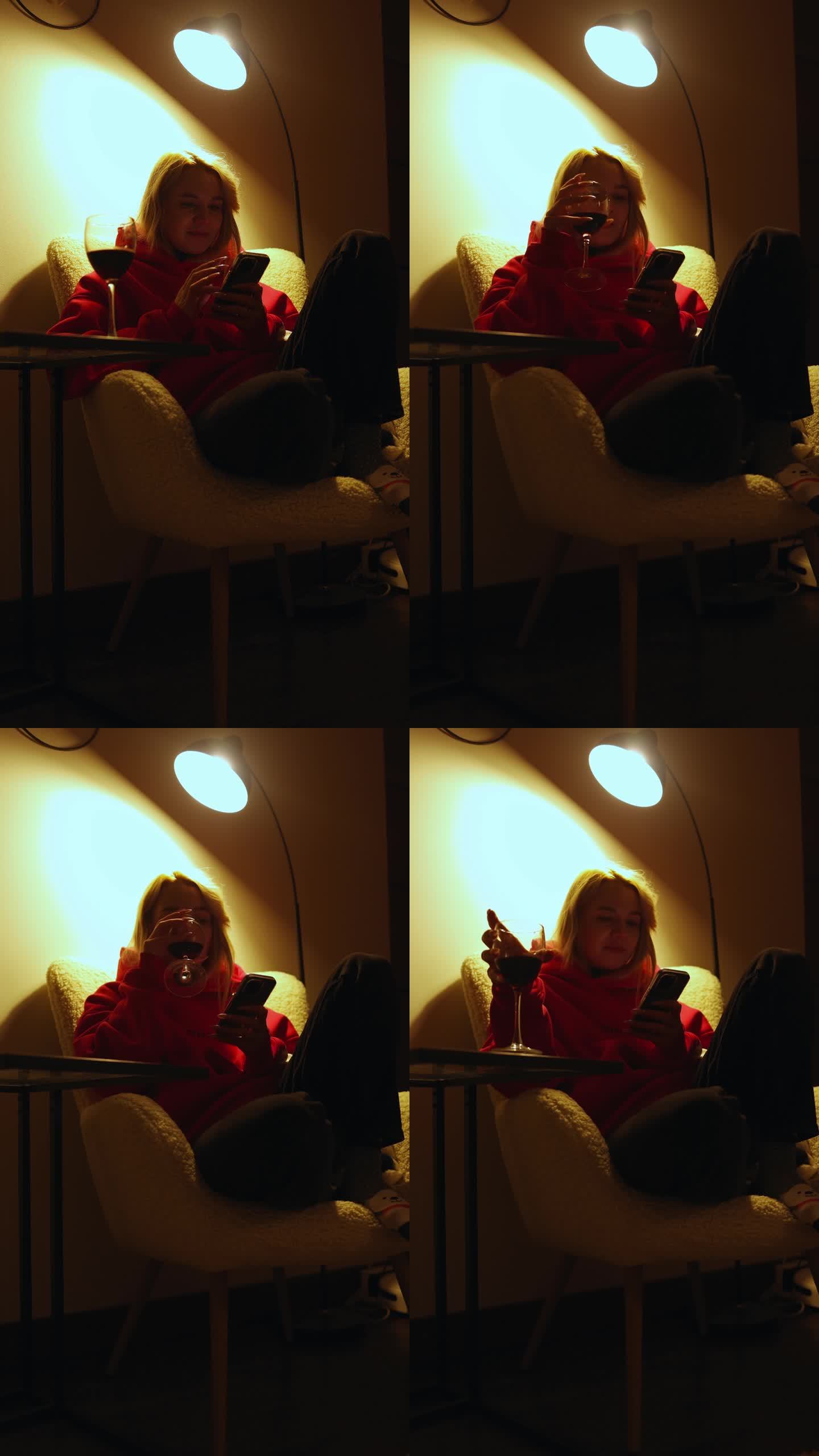 晚上，一个穿着红毛衣的漂亮女孩坐在落地灯下的椅子上，喝着酒，看着手机。垂直视频