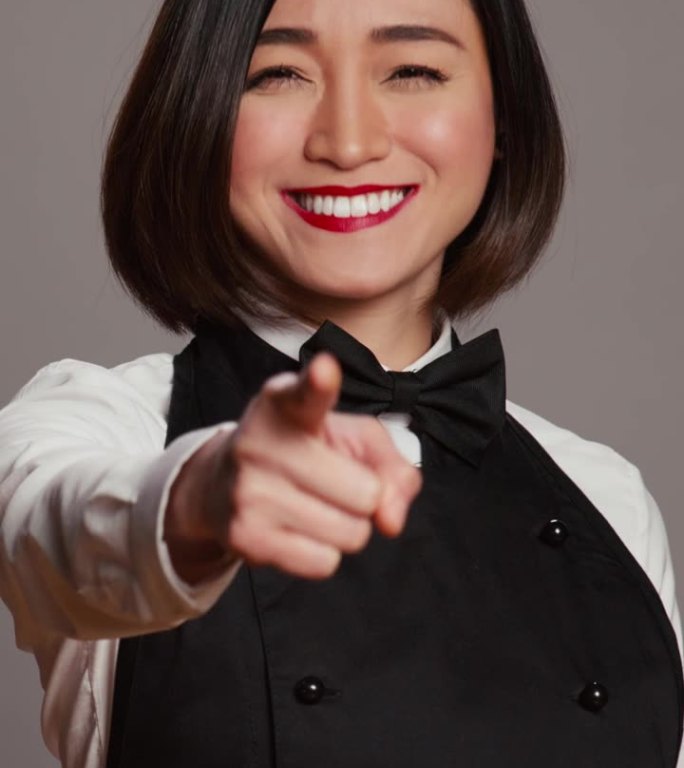 垂直视频亚洲女服务员指着镜头在灰色背景中选择你
