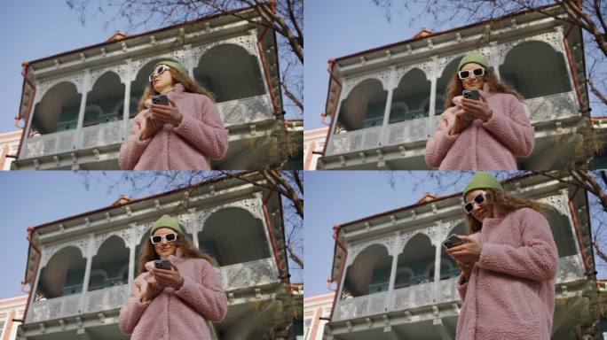 一个时髦的女人站在那里，手里拿着手机，读着有趣的新闻，或者戴着太阳镜在街上与朋友在线交流