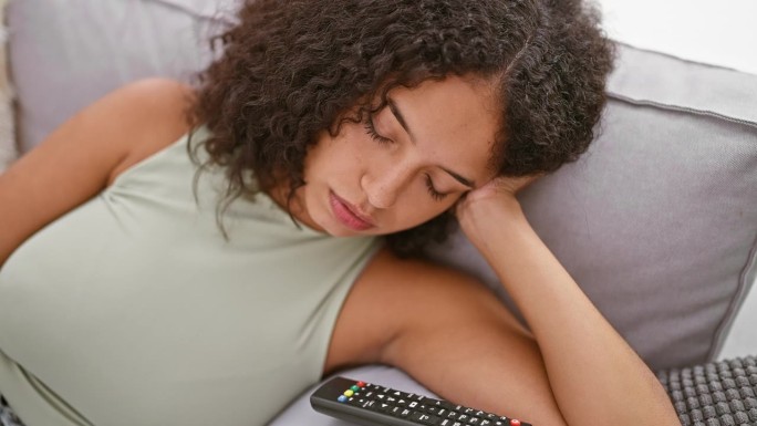 筋疲力尽的年轻西班牙女子躺在沙发上，在家里客厅舒适的背景下睡得很熟，把遥控器留给了急需休息的人