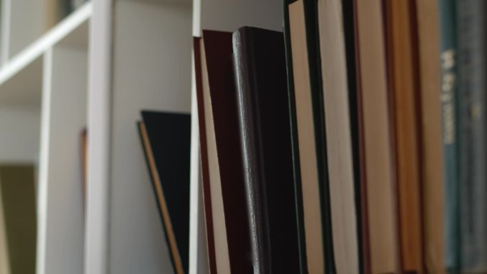 一只手小心翼翼地从家里白色书柜的书架上取出一本书的特写。好奇心和求知欲的概念