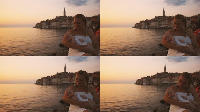 日落时分，快乐的母亲坐在海边的岩石上抱着孩子。罗维尼，伊斯特拉，克罗地亚。