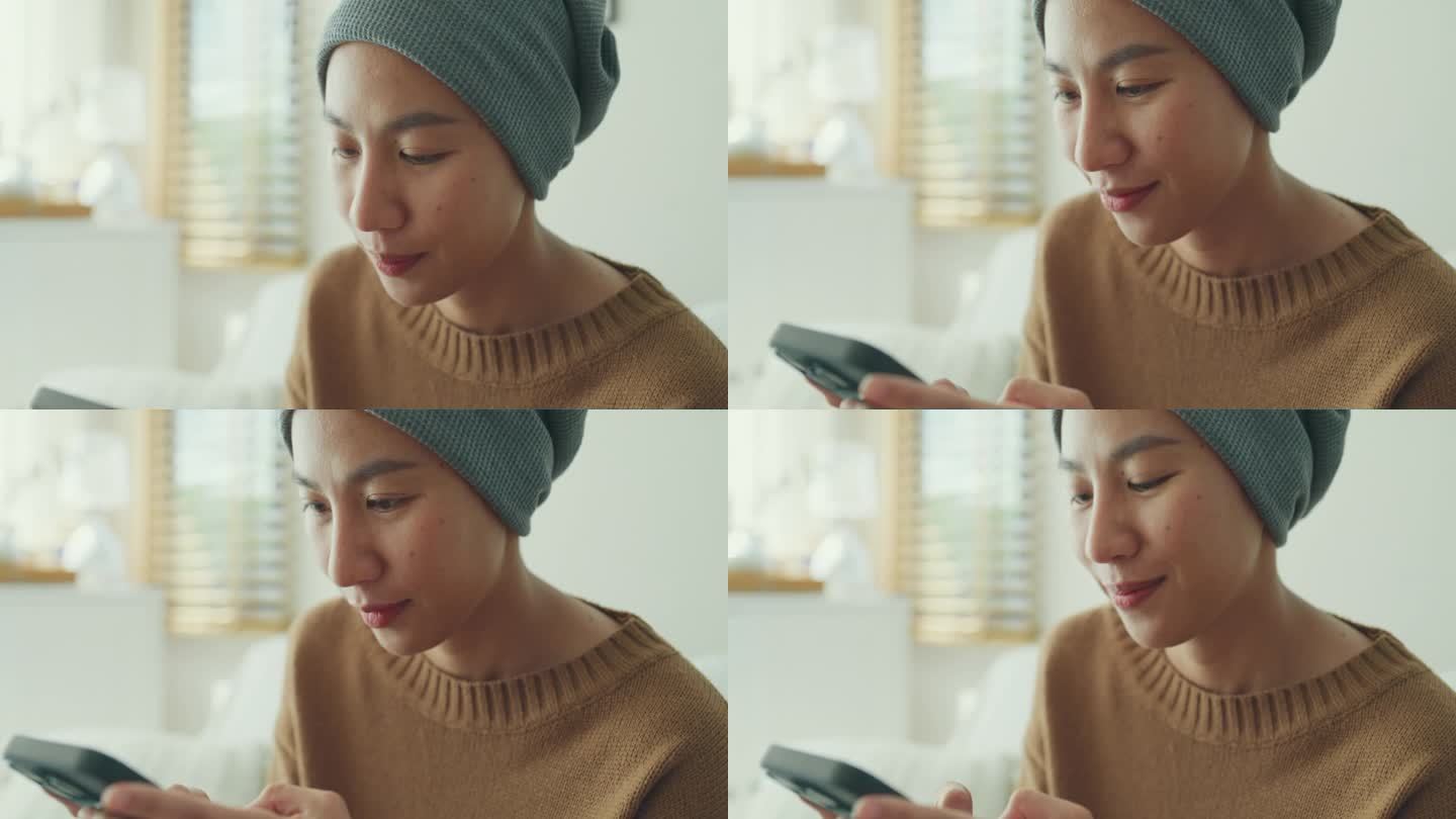 一个患癌症的年轻亚洲女人的特写，她穿着便服，戴着头巾，坐在舒适的沙发上玩手机，周围是一个明亮的，充满