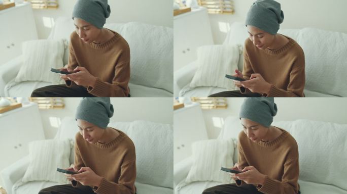 一位年轻的亚洲癌症患者穿着便服，戴着头巾，坐在舒适的沙发上玩手机，周围是一个明亮的，充满植物的客厅。