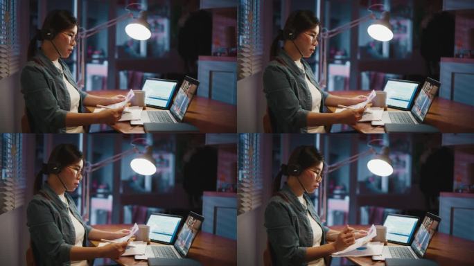 年轻的亚洲女性坐在黑暗的客厅里，戴着耳机在笔记本电脑上进行视频会议