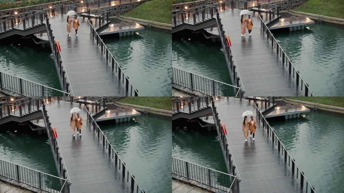 雨中在桥上散步的女人，在大自然中亲密接触，一起度假或周末。聊着天，湖边的朋友或年轻人打着伞在冬天的木