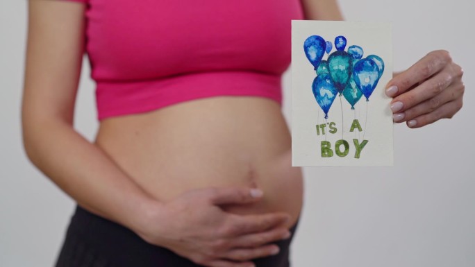 特写镜头。一位孕妇发现了她未出生的孩子的性别，她手里拿着一幅画。一个无法辨认的女人站在白色的背景上。