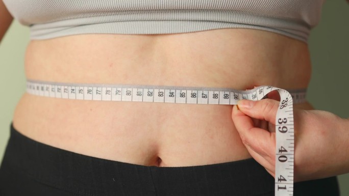 超重女性用卷尺测量腹部脂肪。胖女拿着卷尺，她用手去挤多余的脂肪，让胖女大吃一惊。