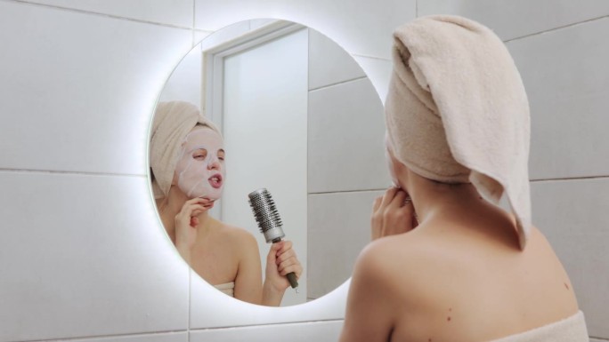有趣的白人妇女，头上裹着毛巾，脸上戴着布面具，拿着梳子，唱歌，在做家庭美容手术时看着镜子，看着浴室里