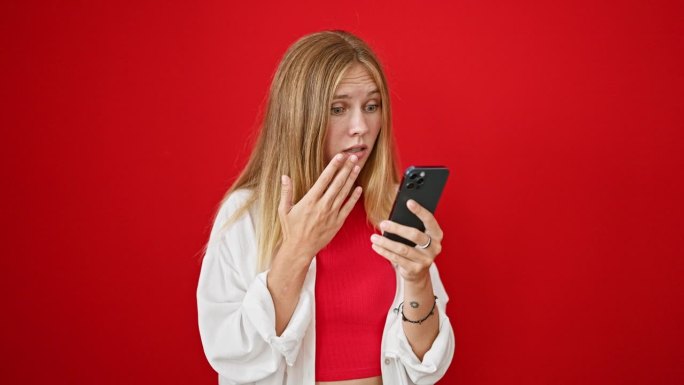 一位身穿白衣的年轻女子惊讶地看着红色背景下的智能手机