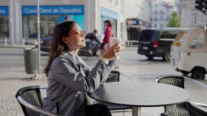 年轻女子坐在街边咖啡馆夏日露台的桌边，端着一杯纸杯咖啡