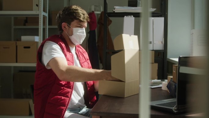 穿着红色背心，戴着防护面具的男子，仓库员工，坐在纸箱的背景上，将包裹装入投递箱并关闭。物流、配送和仓