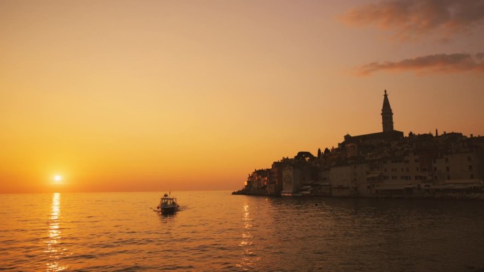 日落时以老城为背景的船在海上航行的慢镜头镜头。罗维尼，伊斯特拉，克罗地亚。