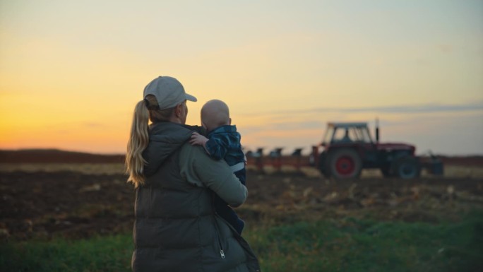 带着小男孩的女农民在夕阳下看着拖拉机在天空中犁地。一位农夫和她的儿子在田里。