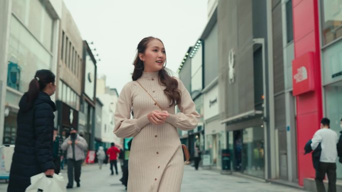 迷人的年轻漂亮的女性穿着休闲的衣服，在街上使用智能手机，与身后的人群快乐地享受韩国济州岛的购物街