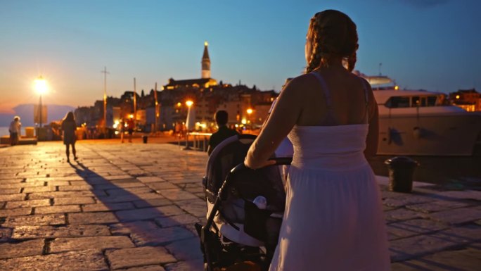 假期黄昏时分，女子推着婴儿车走在灯火通明的小镇上。罗维尼，伊斯特拉，克罗地亚。